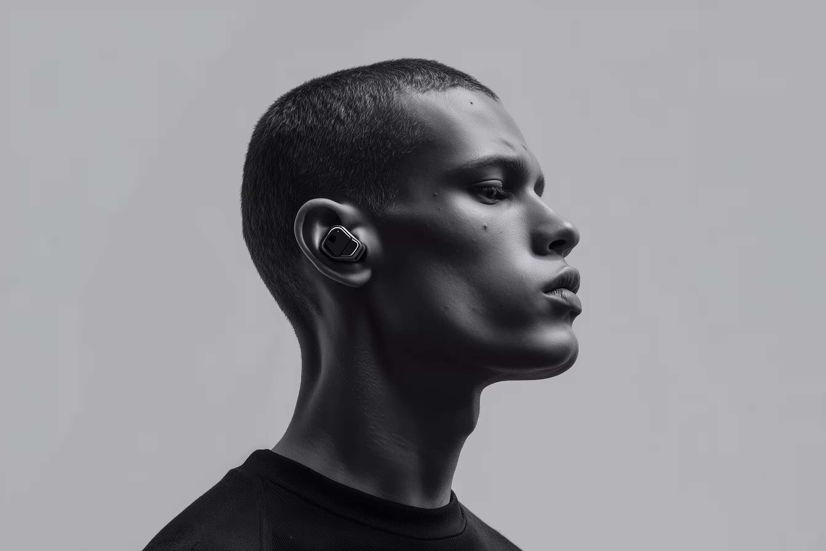 Profile of a young man wearing black wireless earbuds against a plain gray background.