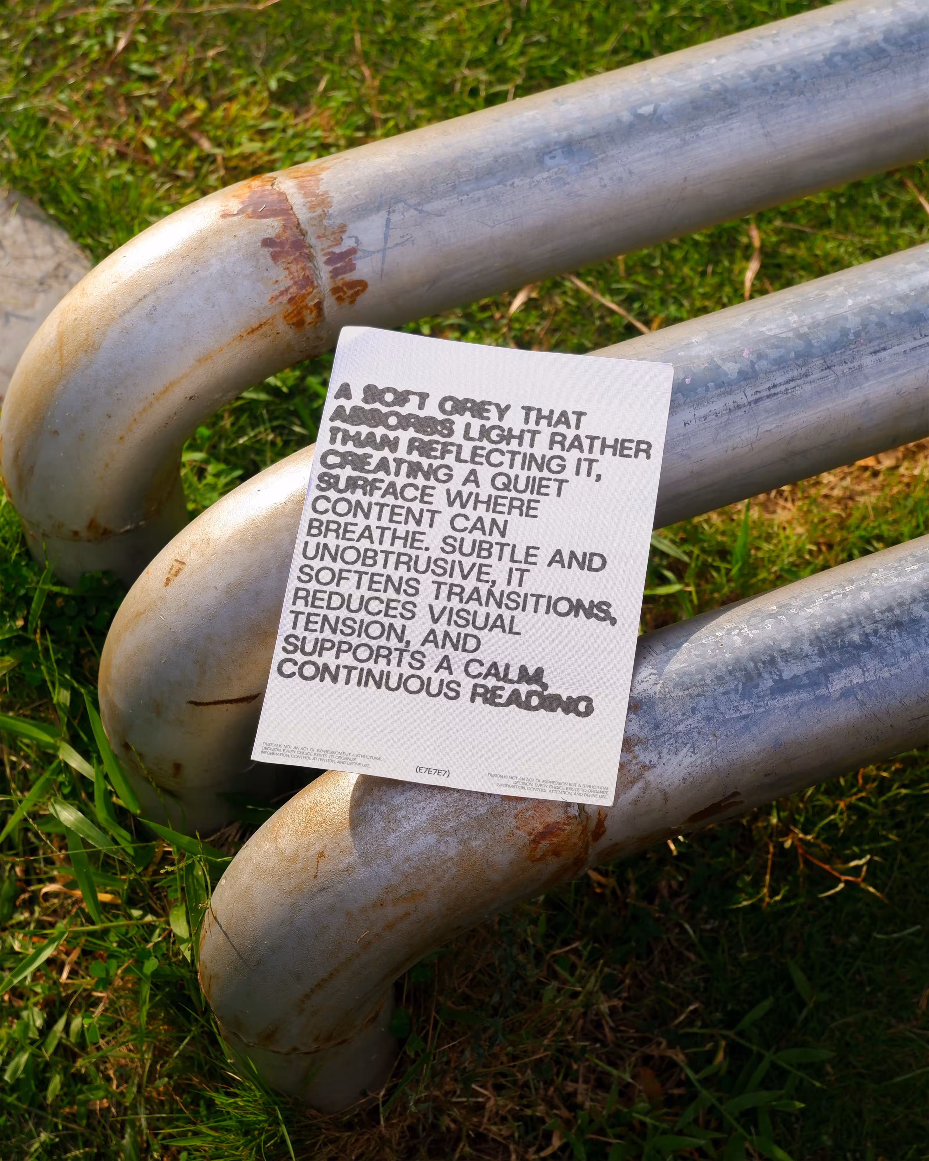 Paper with design text rests on curved metal pipes on grass in sunlight.